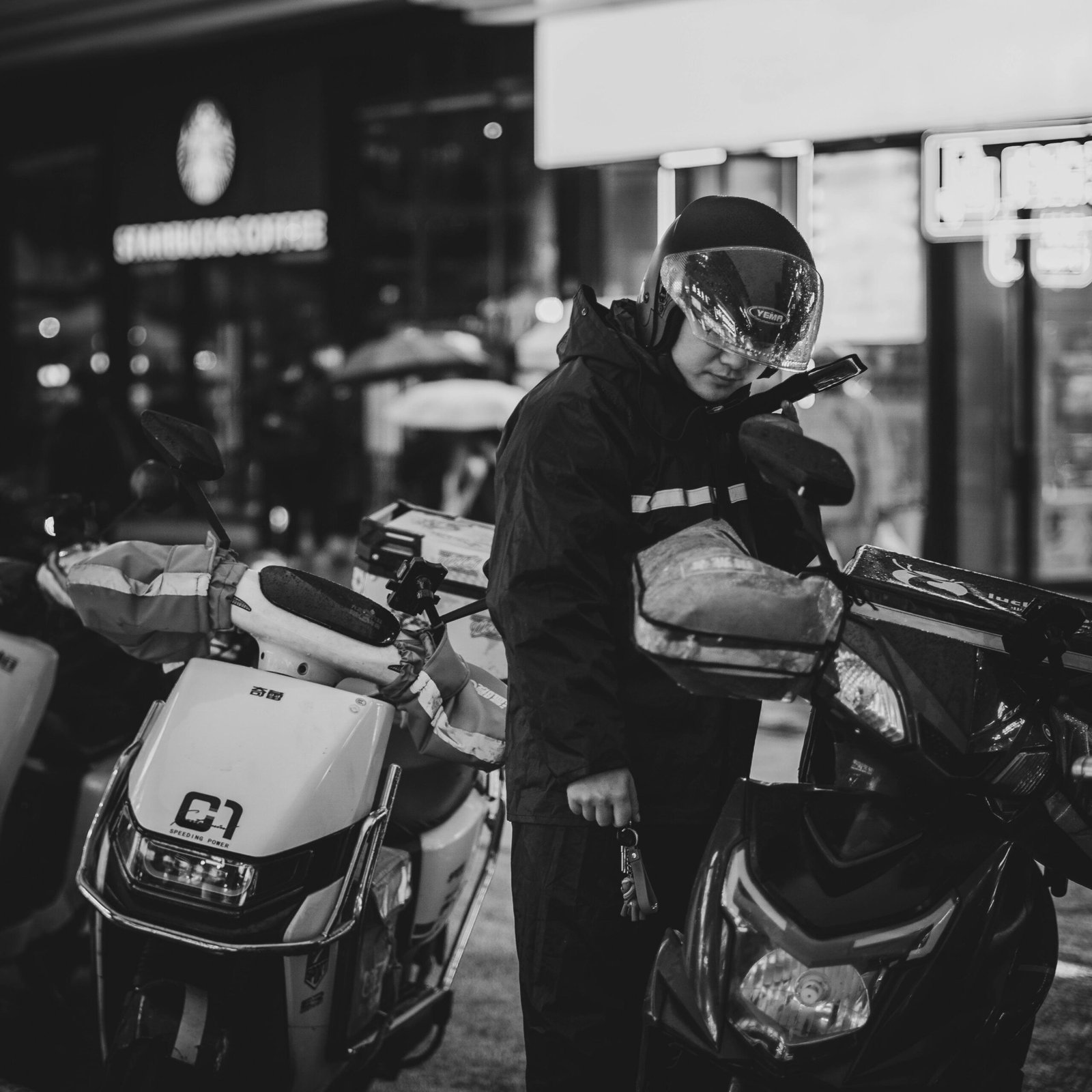 A man wearing a crash helmet stands among parked motor scooters at night in a city.